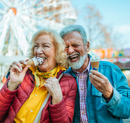 Two people enjoying ice cream cones at a fairground, with a ferris wheel in the background on a bright, sunny day.