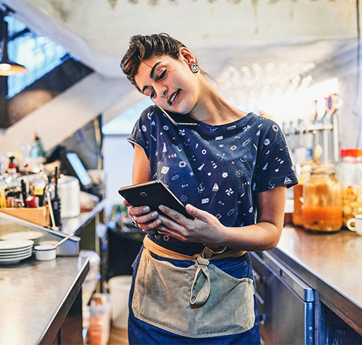 A person wearing a casual printed shirt and an apron is talking on the phone while taking stock on a tablet behind the counter in a cosy café or bar setting.