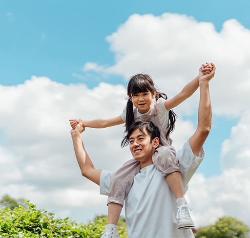 A father is carrying their daughter on their shoulders, holding the child's hands against a backdrop of a bright blue sky with fluffy white clouds.